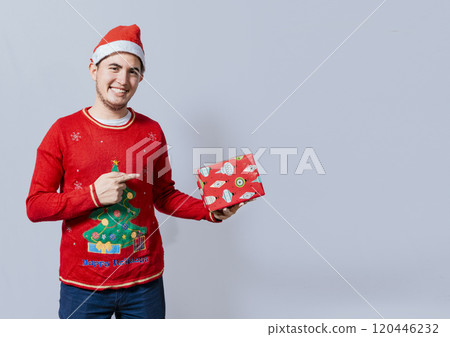 Smiling guy in christmas hat holding and pointing a gift box. Portrait of teenager in christmas hat holding and pointing a gift box. Guy in christmas hat and sweater holding a gift box isolated 120446232