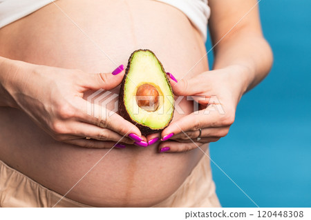 Close-up of a pregnant belly with an avocado held by hands against a blue background 120448308