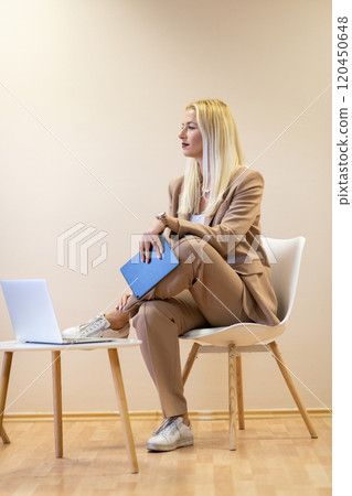 A professional businesswoman sits on a chair, surrounded by a serene beige background, diligently working on her laptop, showcasing dedication and focus in her pursuit of success 120450648