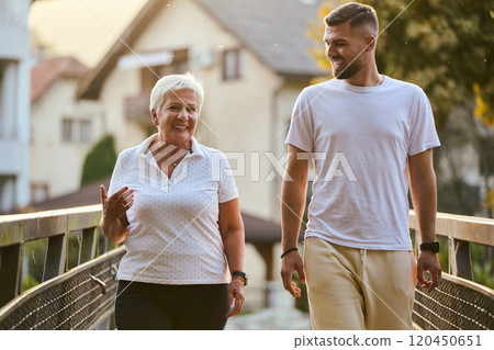 A handsome man and an older woman share a serene walk in nature, crossing a beautiful bridge against the backdrop of a stunning sunset, embodying the concept of a healthy and vibrant intergenerational 120450651