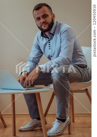 A confident businessman sitting and using laptop with a determined expression, while a beige background enhances the professional atmosphere, showcasing his productivity and expertise. 120450916