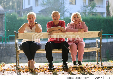 A group of elderly individuals, including a senior man and two older women, sits in a park on a sunny autumn day, embodying the concept of healthy aging through companionship, relaxation, and outdoor 120451004