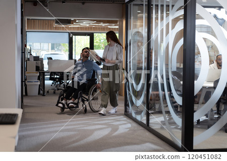 Young business colleagues, collaborative business colleagues, including a person in a wheelchair, walk past a modern glass office corridor, illustrating diversity, teamwork and empowerment in the 120451082