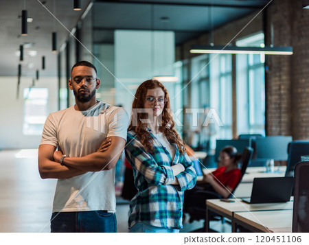 African-American businessman and his businesswoman colleague stand at the forefront with crossed arms, exuding confidence and leadership, while their diverse team diligently works behind them 120451106