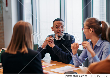 In the vibrant ambiance of a modern startup office, a diverse team of business professionals engage in discussions on various projects and develop new business strategies, epitomizing creativity and 120451107