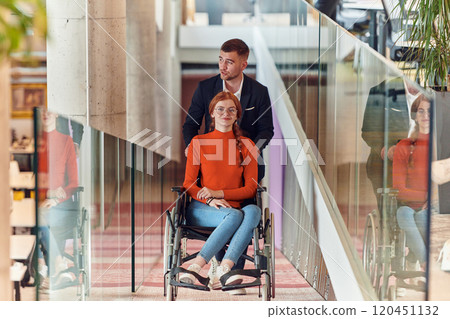 A company director assists his business colleague in a wheelchair, helping her navigate to their startup office, where they work alongside their diverse team of colleagues, emphasizing inclusivity and 120451132