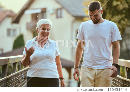 A handsome man and an older woman share a serene walk in nature, crossing a beautiful bridge against the backdrop of a stunning sunset, embodying the concept of a healthy and vibrant intergenerational 120451444
