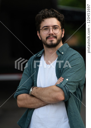 A successful young businessman in a shirt, with crossed arms, poses outdoors, confident expression on his face. A successful young businessman in a shirt, with crossed arms, poses outdoors, confident expression on his face. 120451697