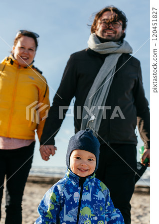 A happy family strolls along a chilly autumn beach, with their young son joyfully running ahead, creating lasting memories as the crisp wind and soothing waves embrace them in a moment of pure 120451827