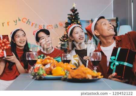 Group of young Asian man and women as friends having fun at a New Year's celebration, holding gift boxes standing by Christmas tree decoration, midnight countdown Party at home with holiday season. 120452122