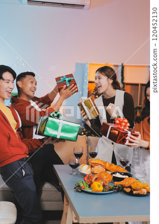 Group of young Asian man and women as friends having fun at a New Year's celebration, holding gift boxes standing by Christmas tree decoration, midnight countdown Party at home with holiday season. 120452130