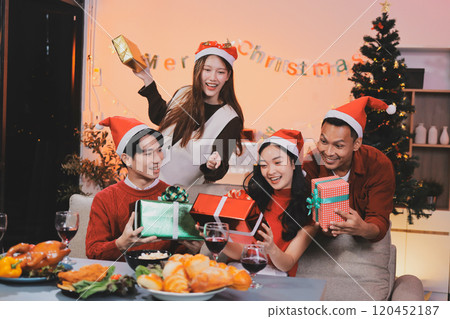 Group of young Asian man and women as friends having fun at a New Year's celebration, holding gift boxes standing by Christmas tree decoration, midnight countdown Party at home with holiday season. 120452187