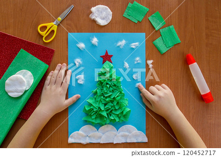 little child making winter decoration "Christmas Tree" from paper, crumpled paper or napkins, and cotton disc. Children's art project. DIY concept. little child making winter decoration "Christmas Tree" from paper, crumpled paper or napkins, and cotton disc. Children's art project. DIY concept. 120452717