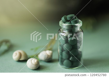 Spirulina pills in glass bottle, sea shells on green background. Antioxidant nutrition. Bio-energy, biofuel, energy research. Copy space Spirulina pills in glass bottle, sea shells on green background. Antioxidant nutrition. Bio-energy, biofuel, energy research. Copy space 120452780
