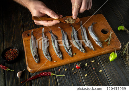 A skilled hand carefully seasons fresh fish laid out on a rustic wooden board, using a wooden spoon to sprinkle aromatic spices. The warm kitchen invites creativity in cooking. A skilled hand carefully seasons fresh fish laid out on a rustic wooden board, using a wooden spoon to sprinkle aromatic spices. The warm kitchen invites creativity in cooking. 120455969