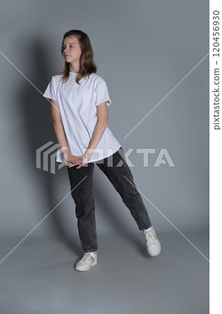 A young girl leans slightly to one side, her hands gently clasped, dressed in casual jeans and a white t-shirt. The minimalist background emphasizes her calm and carefree expression. 120456930