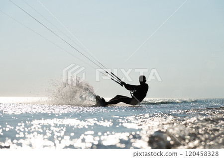 Kite surfing on waves. Silhouette of athletic man riding fast on kiteboard in sea, creating waves and splashes on water Kite surfing on waves. Silhouette of athletic man riding fast on kiteboard in sea, creating waves and splashes on water 120458328