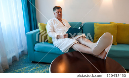 A man in a robe sits comfortably on a sofa with his feet on the table, choosing a menu for breakfast at a hotel A man in a robe sits comfortably on a sofa with his feet on the table, choosing a menu for breakfast at a hotel 120458460