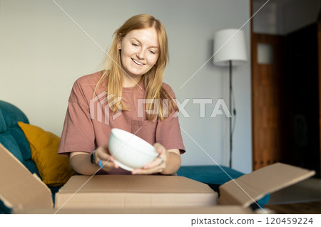 Young 30s woman opening parcel with plates indoors. Happy millennial female client satisfied with ordered purchase. Fast delivery service, online shopping concept Young 30s woman opening parcel with plates indoors. Happy millennial female client satisfied with ordered purchase. Fast delivery service, online shopping concept 120459224