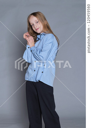 Dressed in a light blue shirt and black pants, a young girl with long blonde hair poses with a gentle smile and a slight head tilt. This image highlights the sweet and approachable nature of childhood 120459560