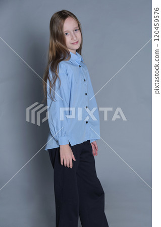 Wearing a light blue shirt and black pants, a young girl stands with a slight head tilt and a soft smile. This image captures the innocence and warmth of childhood, highlighting her friendly demeanor. Wearing a light blue shirt and black pants, a young girl stands with a slight head tilt and a soft smile. This image captures the innocence and warmth of childhood, highlighting her friendly demeanor. 120459576