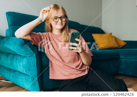 Beautiful Caucasian pleased woman smiling and using cellphone while sitting near sofa on floor in apartment. Phone Communication and comfort concept Beautiful Caucasian pleased woman smiling and using cellphone while sitting near sofa on floor in apartment. Phone Communication and comfort concept 120462428