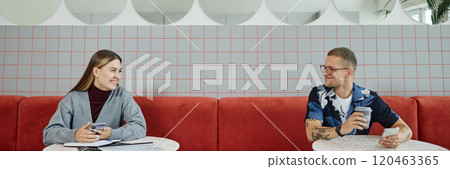Website header shot of cheerful businesswoman in conversation with male colleague sitting at tables during coffee break at cafe featuring modern minimalistic design with red elements, copy space 120463365