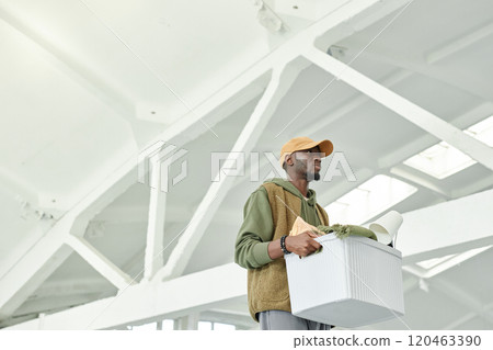 Medium shot of Black man carrying box with personal belongings towards rented self storage unit captured against high warehouse ceiling while moving out from apartment, copy space 120463390