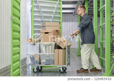 Full length shot of handsome man loading cart with cardboard boxes into self storage unit at warehouse while moving out or decluttering apartment 120463410