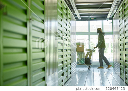 Side view of Black man pushing cart loaded with cardboard boxes full of home items walking along warehouse hallway towards self storage unit against window while moving out, copy space Side view of Black man pushing cart loaded with cardboard boxes full of home items walking along warehouse hallway towards self storage unit against window while moving out, copy space 120463423