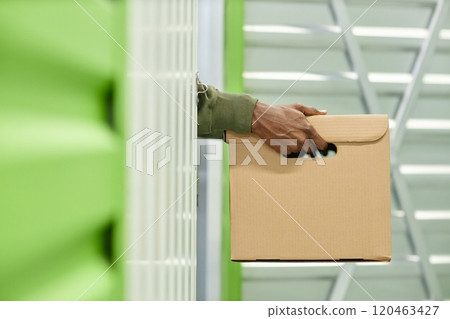 Close up on hands of Black man carrying cardboard box filled with personal belongings loading home stuff into self storage unit at warehouse providing convenient temporary service, copy space Close up on hands of Black man carrying cardboard box filled with personal belongings loading home stuff into self storage unit at warehouse providing convenient temporary service, copy space 120463427