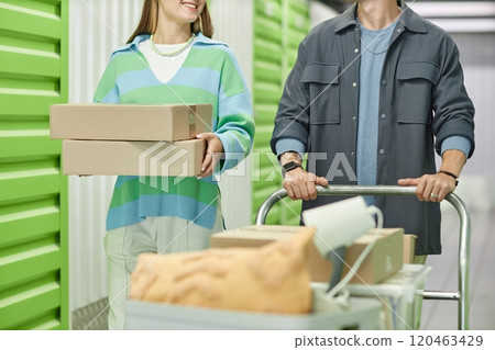 Cropped shot of smiling woman carrying pile of cardboard boxes walking next to boyfriend helping with moving out, while pushing loaded dolly cart in hallway of self storage warehouse 120463429