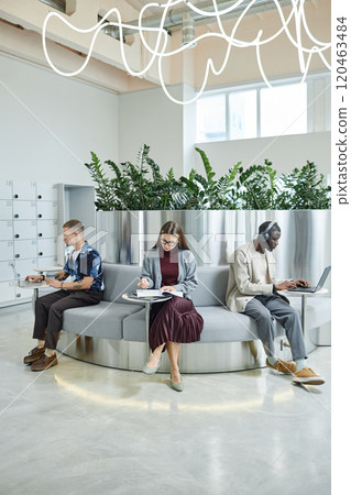 Vertical shot of businesswoman writing in notebook while sitting at table next to coworkers in designer lounge area featuring biophilic design with metallic elements, copy space 120463484