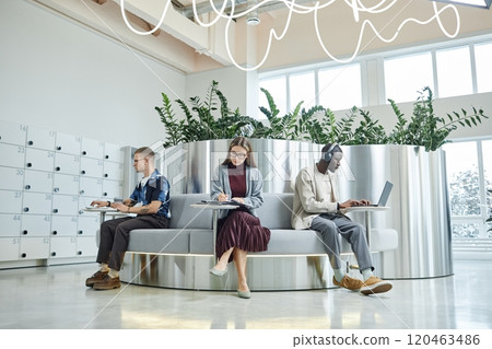 Wide angle shot of businesswoman writing in notebook while sitting at table next to colleagues in designer lounge zone featuring biophilic design and metallic elements 120463486