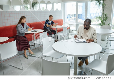Wide angle shot of African American businessman writing in notebook with business people checking phones in background, while sitting at tables in cozy cafe featuring modern minimalistic design 120463537