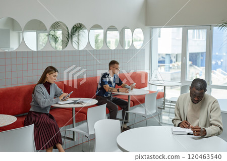 Wide angle shot of African American businessman writing in notebook while sitting at table in cafe of coworking space. Business people working in cozy cafe featuring modern minimalistic design Wide angle shot of African American businessman writing in notebook while sitting at table in cafe of coworking space. Business people working in cozy cafe featuring modern minimalistic design 120463540