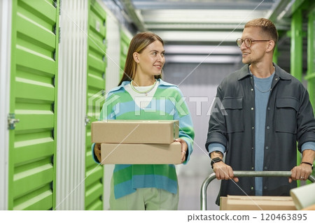 Medium shot of smiling woman carrying cardboard boxes walking along self storage warehouse hallway with helping boyfriend pushing loaded dolly cart Medium shot of smiling woman carrying cardboard boxes walking along self storage warehouse hallway with helping boyfriend pushing loaded dolly cart 120463895