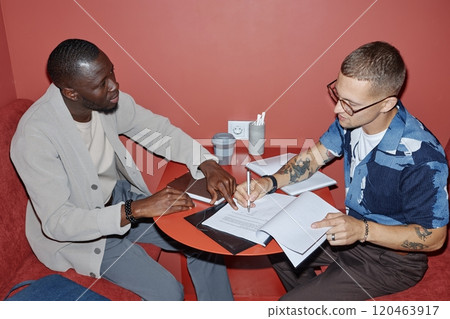Side view of African American financial adviser showing place for signature on document to male client while sitting at table in coworking cafe, camera flash, copy space Side view of African American financial adviser showing place for signature on document to male client while sitting at table in coworking cafe, camera flash, copy space 120463917