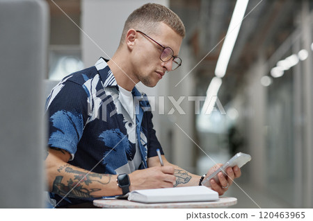 Side view of young male business worker checking message on mobile phone writing in notebook while sitting at table in common area of modern office building, copy space 120463965