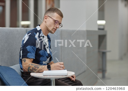 Side view of young male business worker concentrated on writing in notebook sitting at table in common area of modern office building, copy space 120463968
