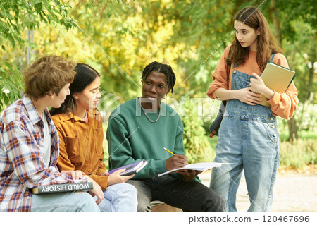 Group of students studying outdoors with diverse and focused expressions, sitting on benches surrounded by greenery and holding books and notebooks Group of students studying outdoors with diverse and focused expressions, sitting on benches surrounded by greenery and holding books and notebooks 120467696