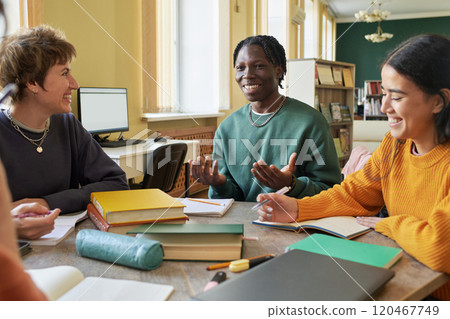 Group of diverse students sitting in classroom engaging in group study session filled with textbooks and notebooks. Natural light streaming through windows brightens room Group of diverse students sitting in classroom engaging in group study session filled with textbooks and notebooks. Natural light streaming through windows brightens room 120467749