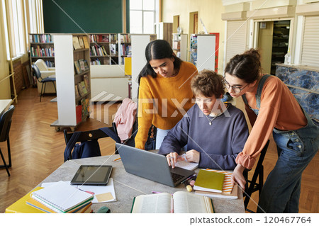 Group of students concentrating on laptop in library setting, with books and notes spread on table. Atmosphere suggests studying and teamwork session without interruptions 120467764