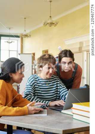 Three young students working together on homework in a bright room. Engaging with each other while using laptop and taking notes with books stacked on table 120467774