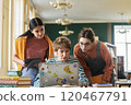 Three students focusing on laptop screen while working on a group project with books and educational tools visible on desk. One of them holding tablet in her hand 120467791