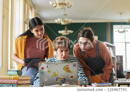 Three students focusing on laptop screen while working on a group project with books and educational tools visible on desk. One of them holding tablet in her hand Three students focusing on laptop screen while working on a group project with books and educational tools visible on desk. One of them holding tablet in her hand 120467791