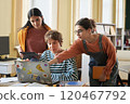 Three students are collaborating on a group project while using a laptop and tablet in classroom environment with bookshelves and windows 120467792