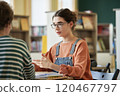 Serious woman engaging in discussion at library study table, focusing on study materials, with bookshelves in background providing academic atmosphere 120467797