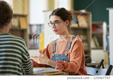 Serious woman engaging in discussion at library study table, focusing on study materials, with bookshelves in background providing academic atmosphere 120467797