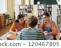 Two young female students sitting at a library table with books, engaging in studying and collaborating on their assignments. Bookshelves with a variety of books in the background 120467805